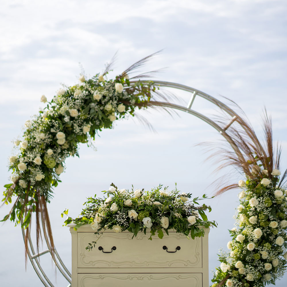 white roses, greenery, baby's breath, and other flowers rest atop stylish boho dresser used as podium and line part of a wedding archway