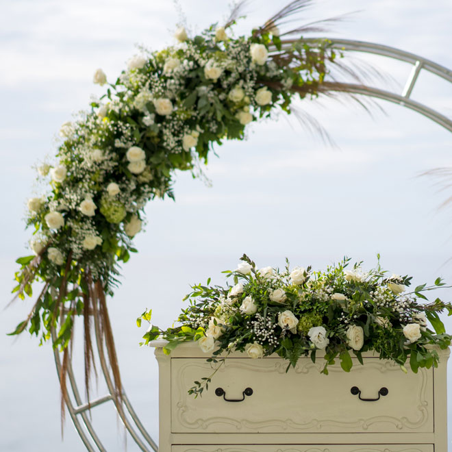 white roses, greenery, baby's breath, and other flowers rest atop stylish boho dresser used as podium and line part of a wedding archway