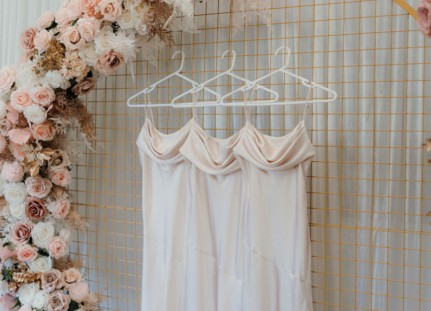 3 silk bridesmaids dresses in champagne hanging beneath a floral arch of pink and white roses