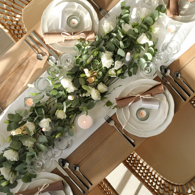 overhead view of a dinner table set with multiple places and a beautiful runner in the center of the table with white roses and greenery. candles are lit throughout